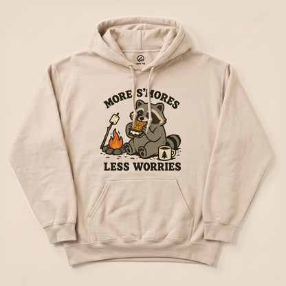 A beige hoodie features a playful graphic of a raccoon enjoying s'mores by a campfire with the text "More S'mores Less Worries.