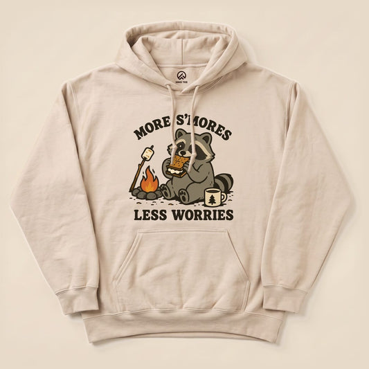 A beige hoodie features a playful graphic of a raccoon enjoying s'mores by a campfire with the text "More S'mores Less Worries.