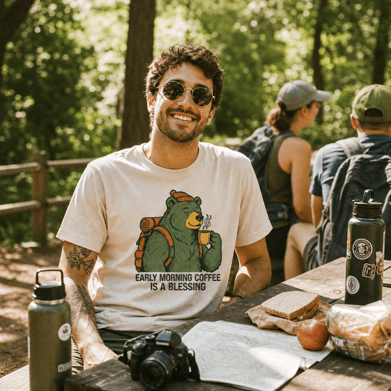 A smiling man sits at a picnic table in the woods wearing a graphic t-shirt featuring a bear holding coffee with the text "Early Morning Coffee Is A Blessing.