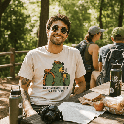 A smiling man sits at a picnic table in the woods wearing a graphic t-shirt featuring a bear holding coffee with the text "Early Morning Coffee Is A Blessing.
