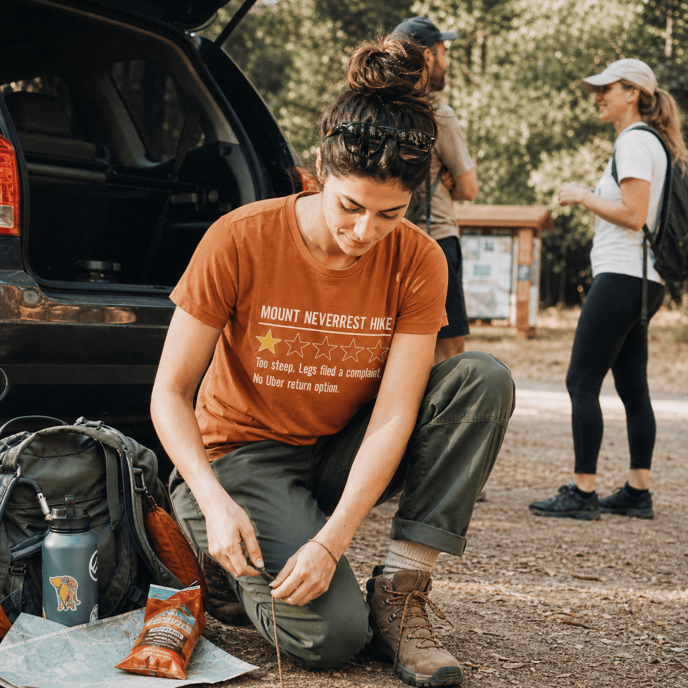 A woman sits on the ground near a car, wearing a rust-colored graphic t-shirt that reads "MOUNT NEVERREST HIKE" with humorous text below.
