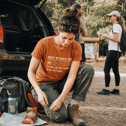 A woman sits on the ground near a car, wearing a rust-colored graphic t-shirt that reads "MOUNT NEVERREST HIKE" with humorous text below.