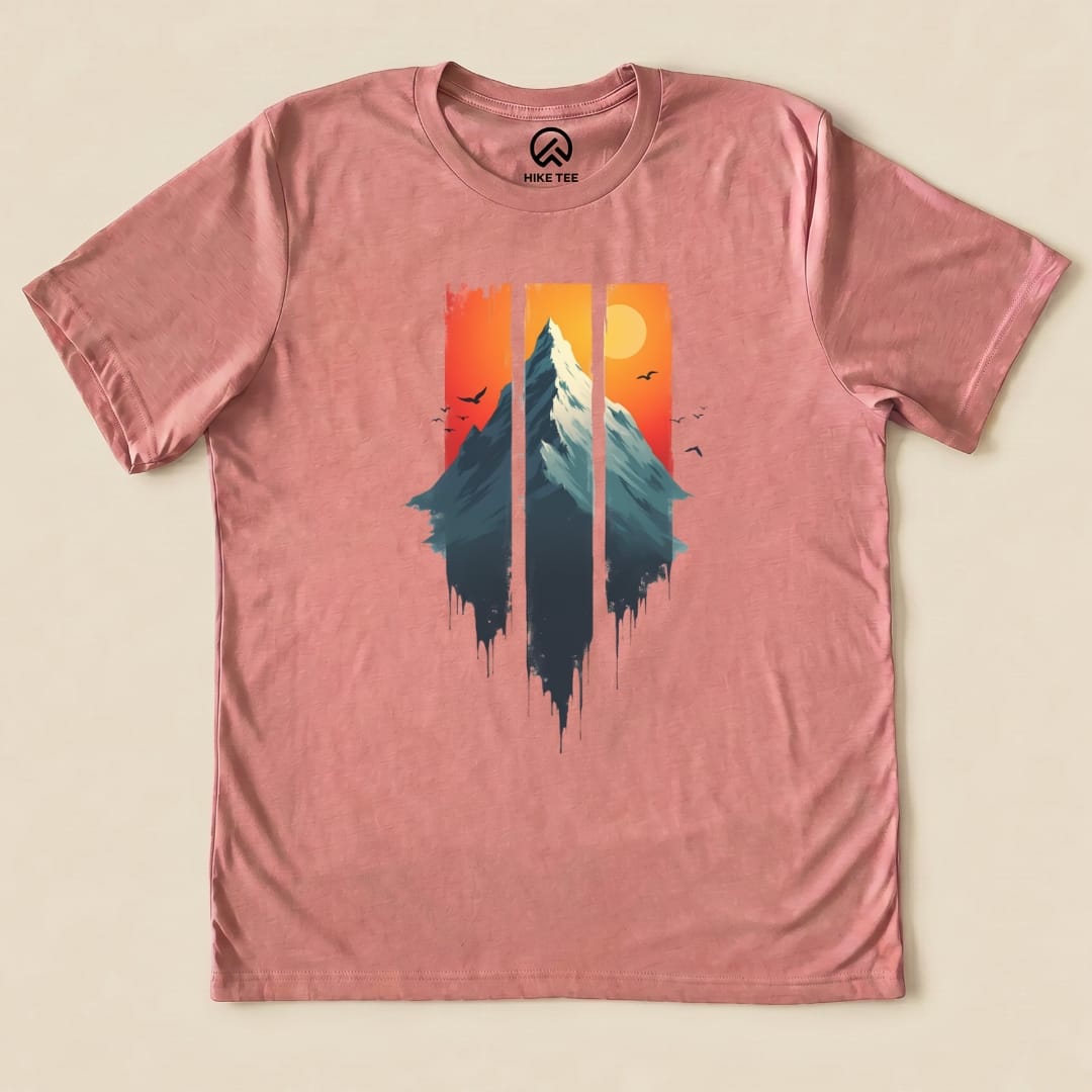A graphic t-shirt in soft pink featuring a mountain silhouette with a sunset background in orange and yellow, titled "Mountain Sunset Shirt".