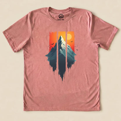 A graphic t-shirt in soft pink featuring a mountain silhouette with a sunset background in orange and yellow, titled "Mountain Sunset Shirt".