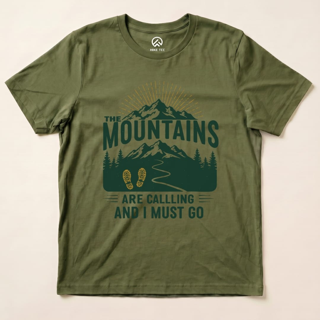 A graphic t-shirt in olive green featuring the phrase "The Mountains Are Calling and I Must Go" with mountain and trail illustrations.