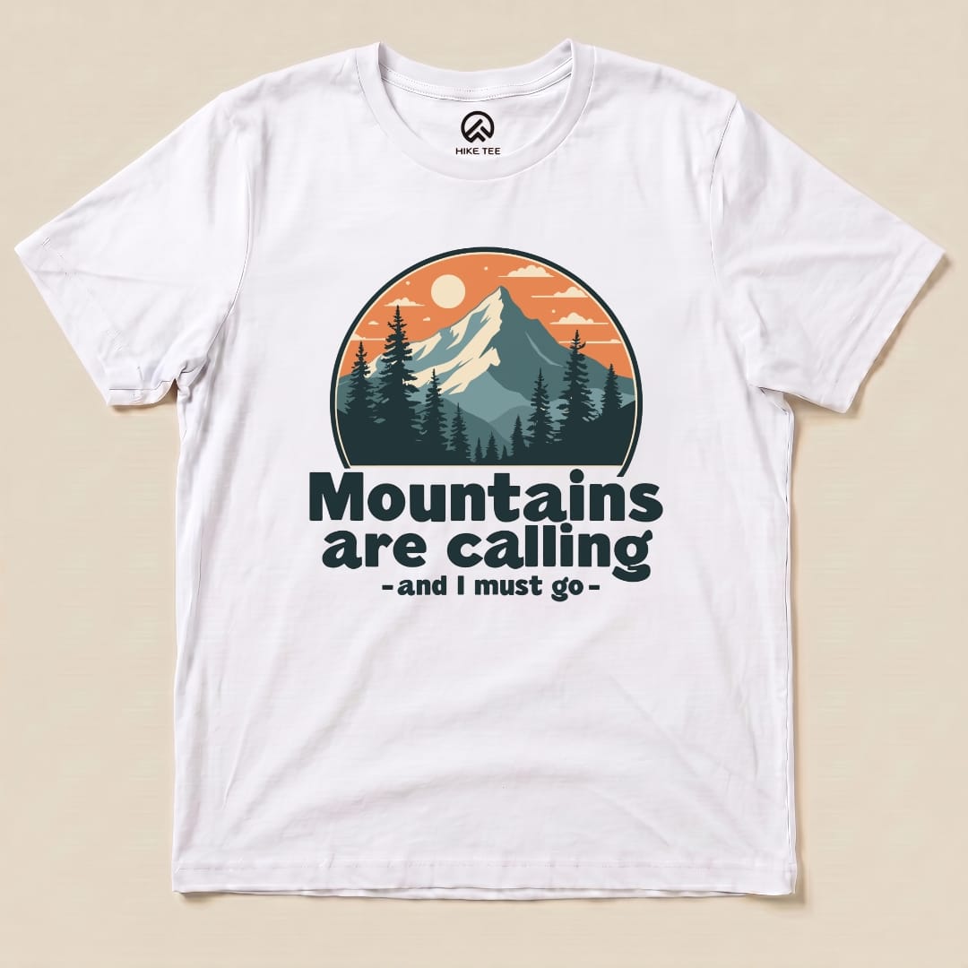 A white graphic t-shirt featuring an illustration of mountains and trees with the text "Mountains are calling - and I must go.