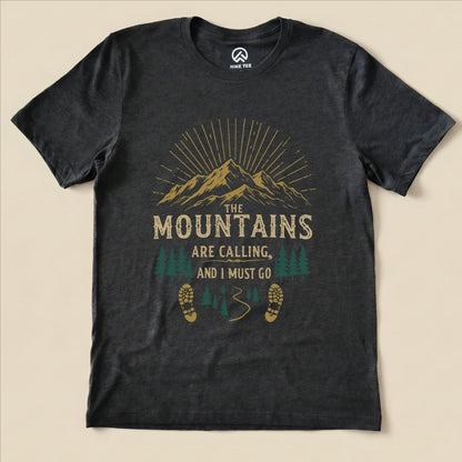 A black graphic t-shirt featuring the phrase "The Mountains are Calling, and I Must Go" with mountains, trees, and footprints.