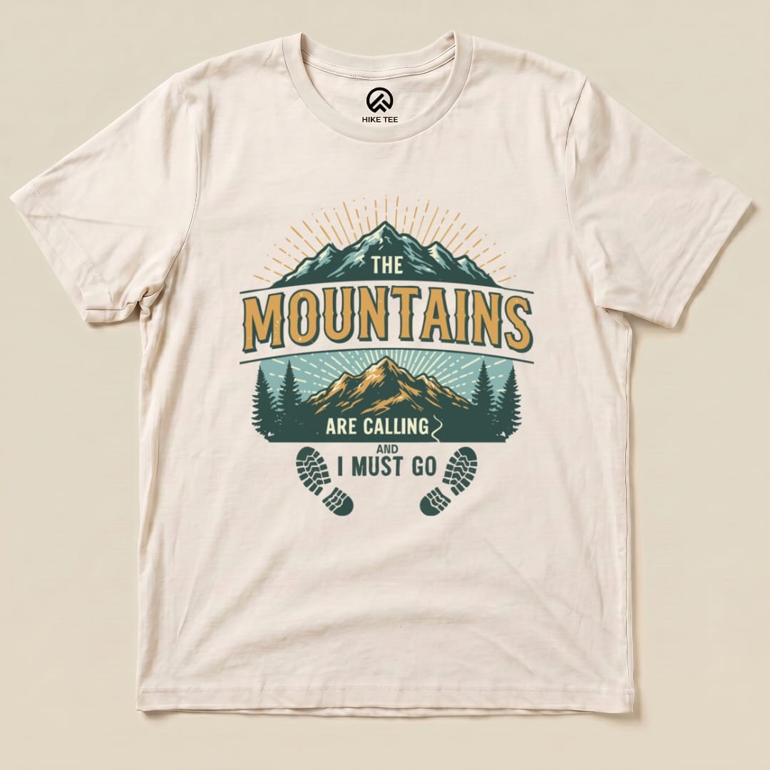 A graphic t-shirt featuring the phrase "THE MOUNTAINS ARE CALLING AND I MUST GO" with mountains, trees, and footprints design.