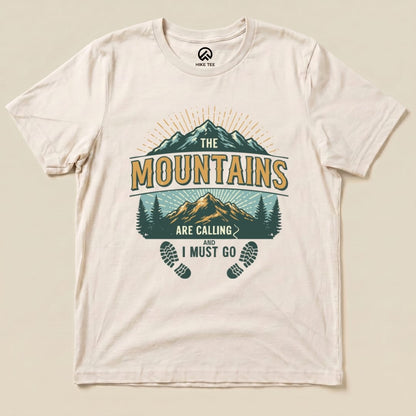 A graphic t-shirt featuring the phrase "THE MOUNTAINS ARE CALLING AND I MUST GO" with mountains, trees, and footprints design.