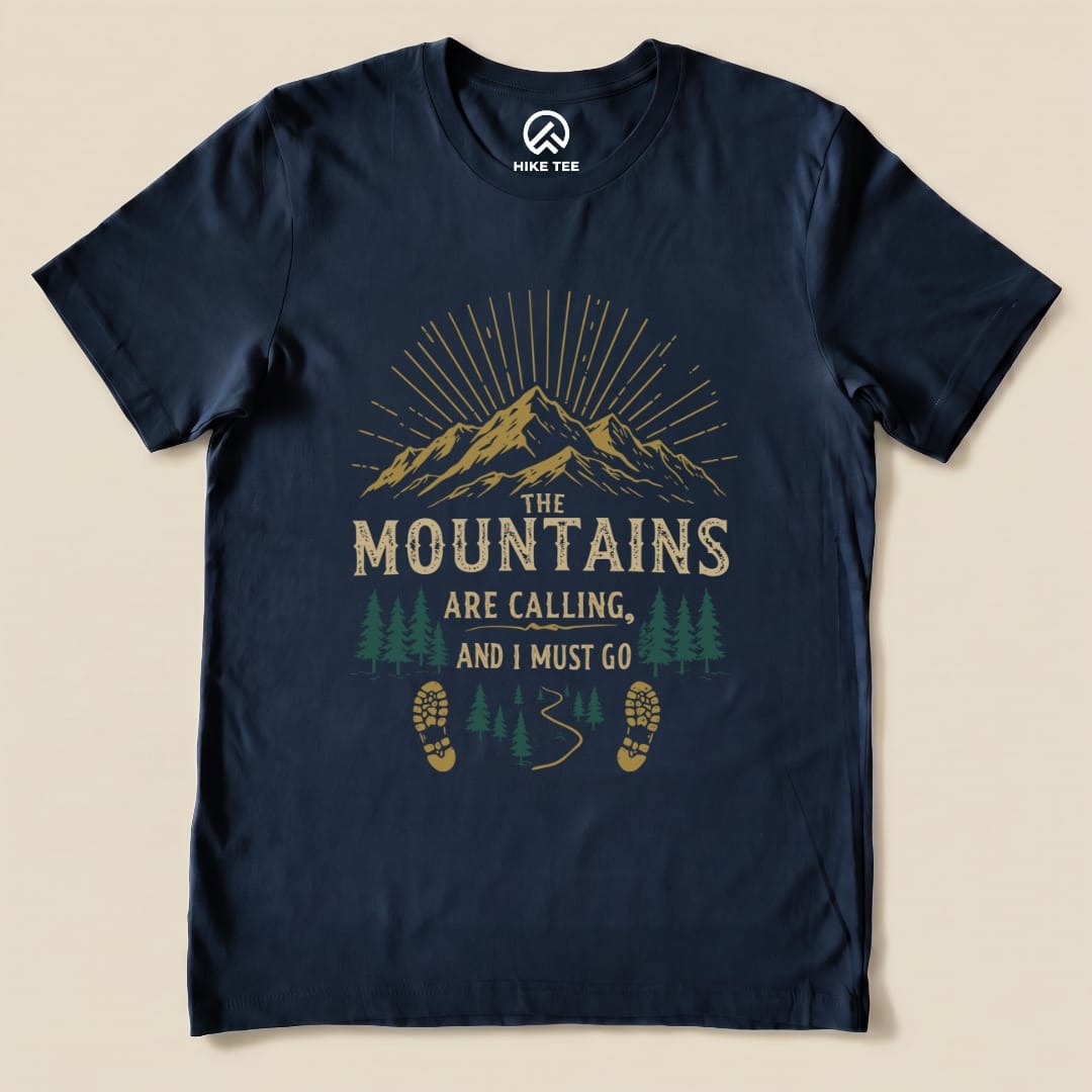 A graphic t-shirt featuring the phrase "The Mountains Are Calling, and I Must Go" with mountains, trees, and footprints design.