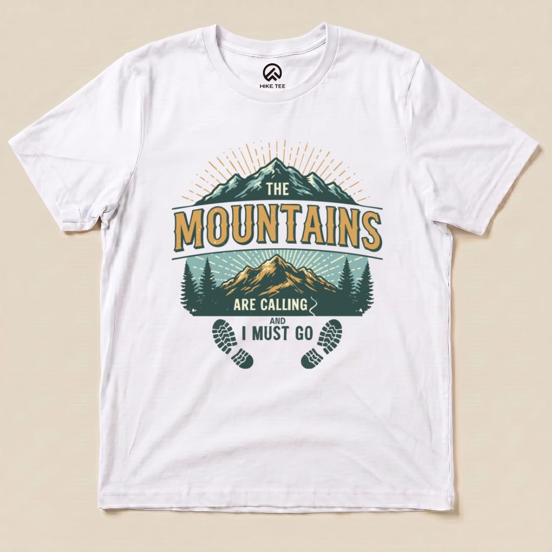 A flat white graphic t-shirt displaying the "Mountains Calling Shirt" design featuring mountains, trees, and the phrase "The Mountains Are Calling and I Must Go.