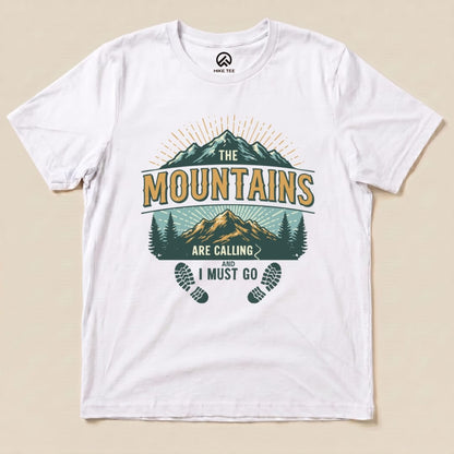 A flat white graphic t-shirt displaying the "Mountains Calling Shirt" design featuring mountains, trees, and the phrase "The Mountains Are Calling and I Must Go.