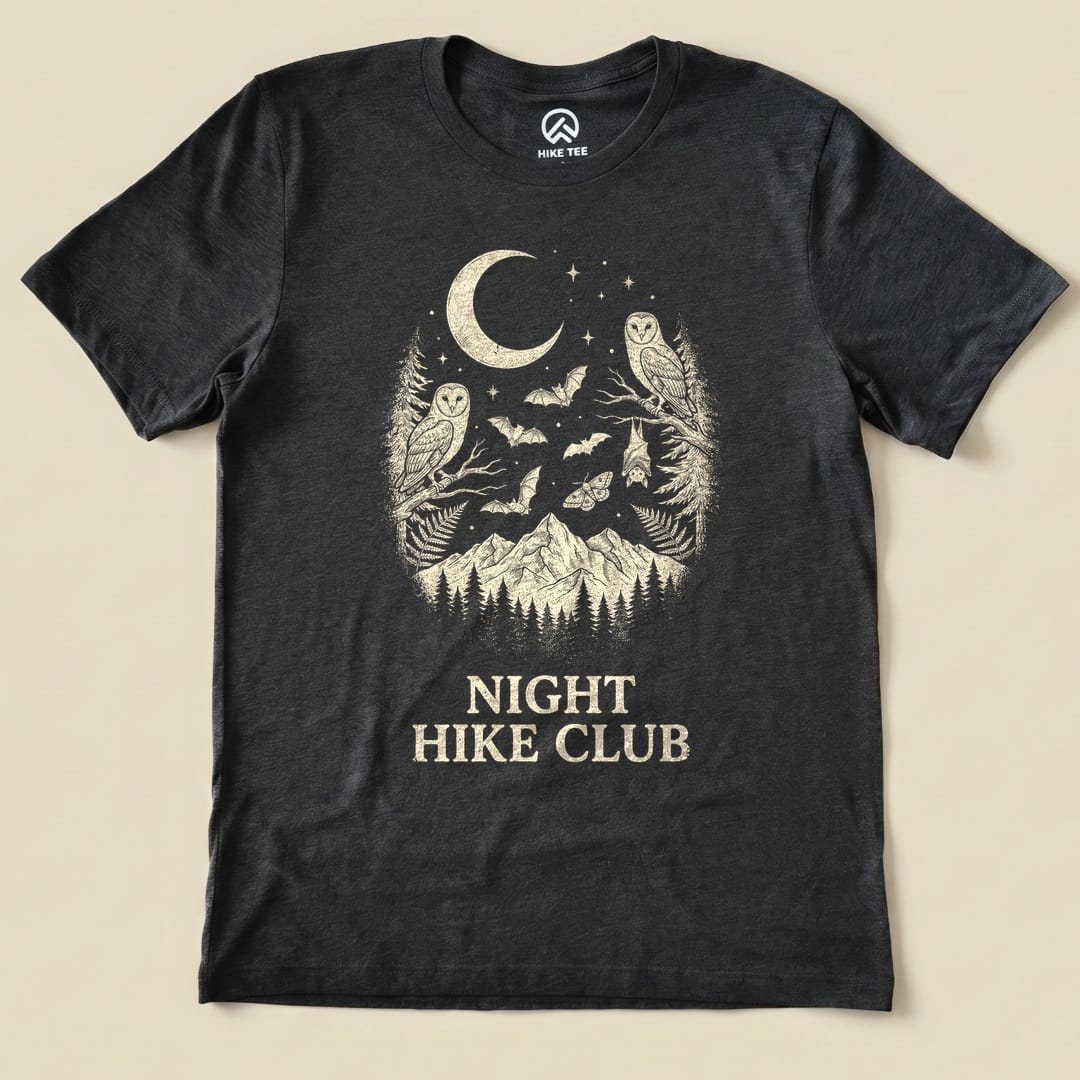 A graphic t-shirt featuring a moon, owls, bats, and mountains, with the phrase "NIGHT HIKE CLUB" printed below.