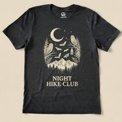 A graphic t-shirt featuring a moon, owls, bats, and mountains, with the phrase "NIGHT HIKE CLUB" printed below.