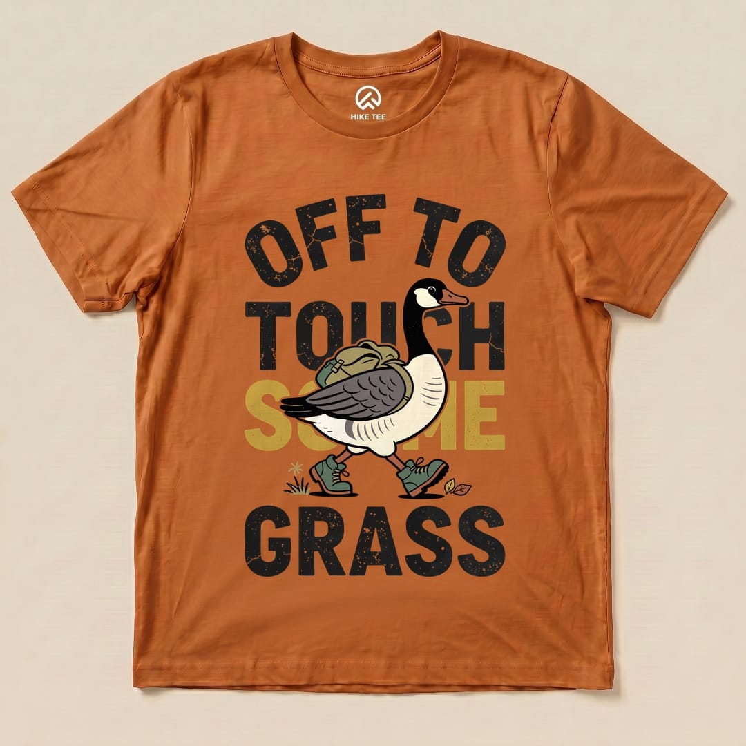 The Hike Tee "Off To Touch Some Grass Shirt" features a cartoon goose walking with a backpack, surrounded by bold text.