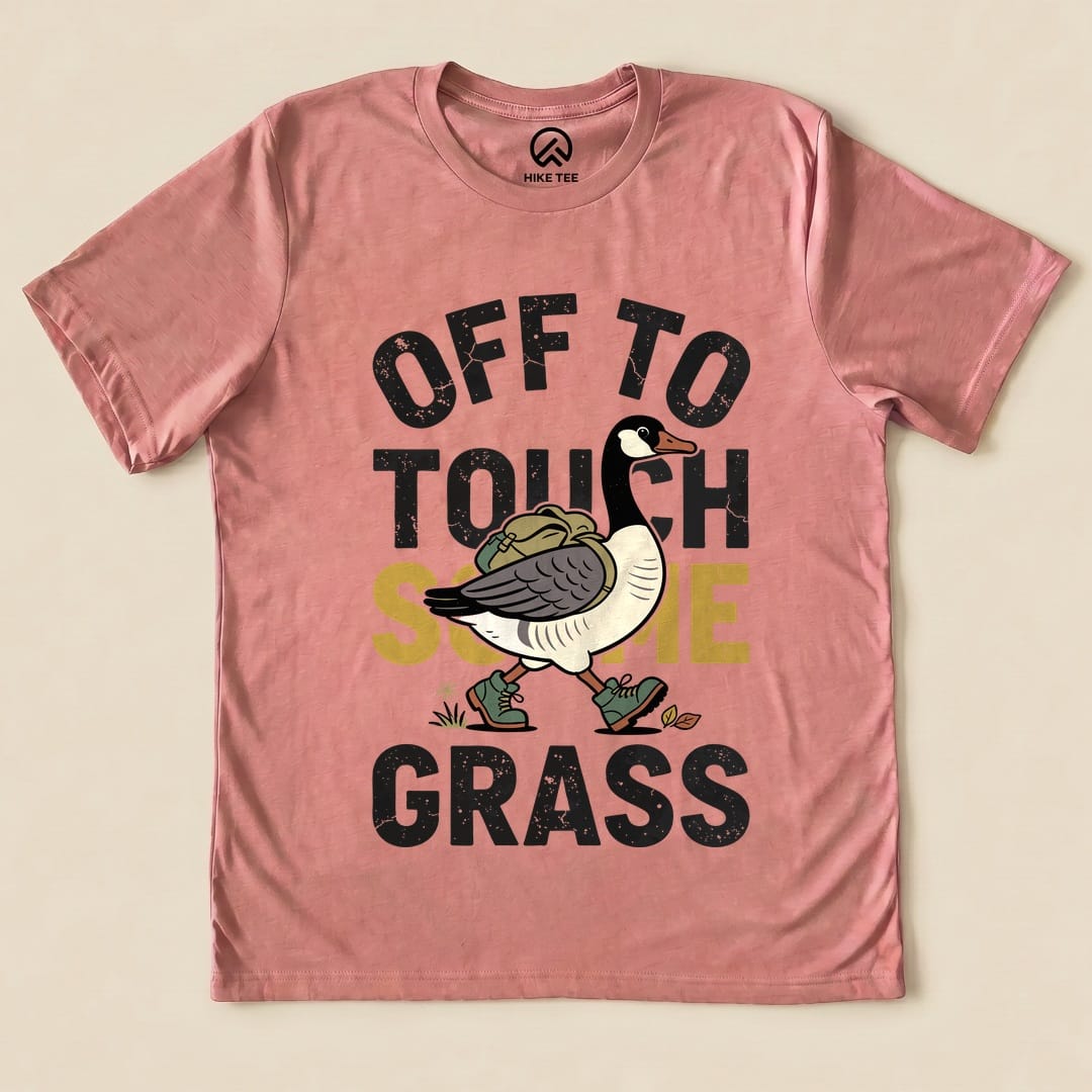 A graphic t-shirt in soft pink featuring a whimsical goose with a backpack and the slogan "Off To Touch Some Grass.
