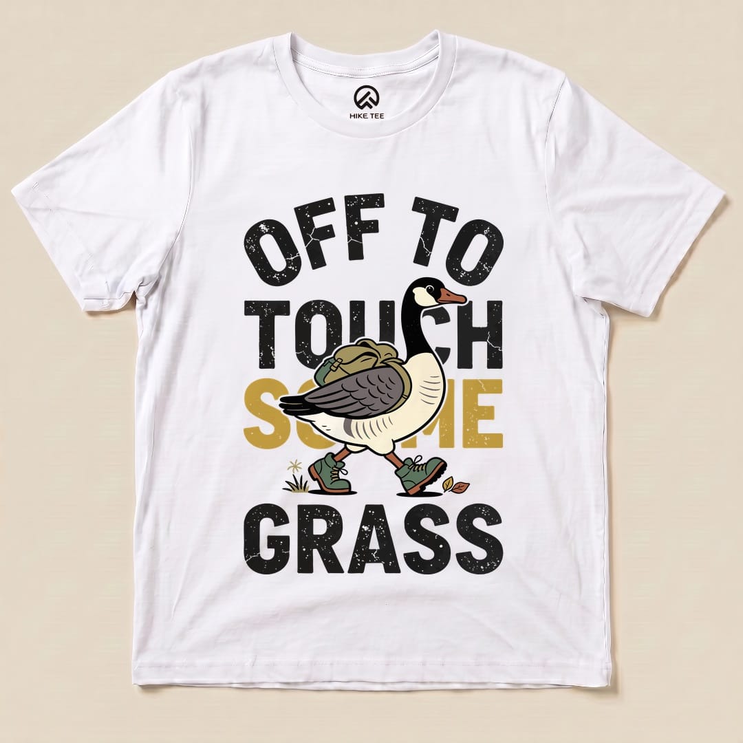 A white graphic t-shirt featuring a cartoon goose with a backpack and the phrase "OFF TO TOUCH SOME GRASS" in bold lettering.