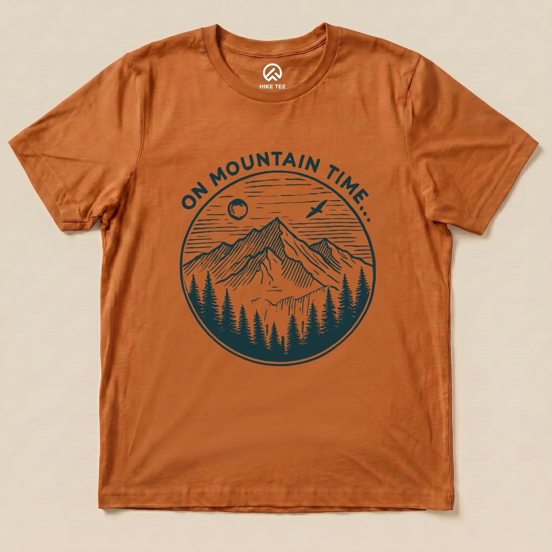 A graphic t-shirt in burnt orange features the phrase "ON MOUNTAIN TIME" above a detailed mountain and forest scene.