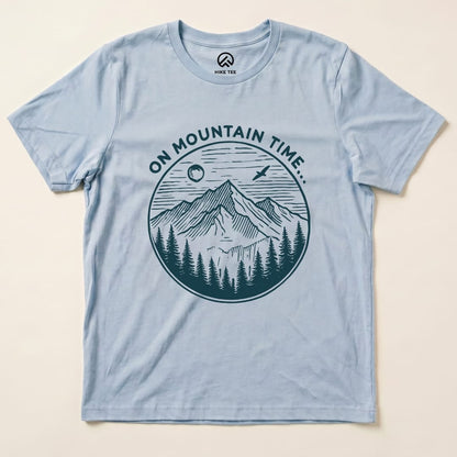 The graphic t-shirt features the phrase "ON MOUNTAIN TIME" above a detailed illustration of mountains, pine trees, and a sun.