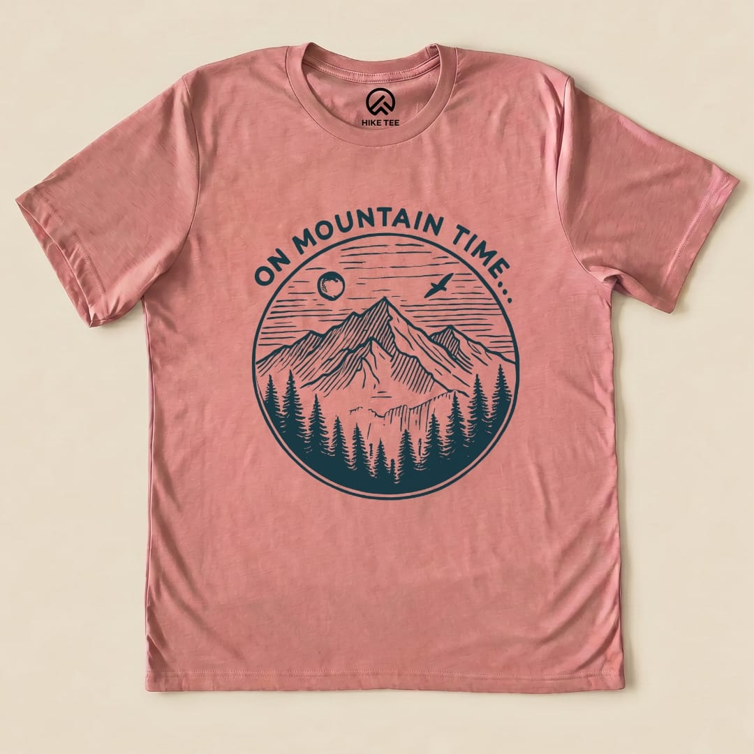 A pink graphic t-shirt featuring the phrase "ON MOUNTAIN TIME..." above a mountain landscape and forest design.