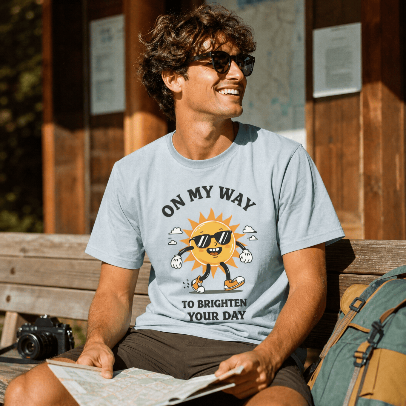 A smiling man sits on a bench outside, wearing a graphic t-shirt that reads "ON MY WAY TO BRIGHTEN YOUR DAY" with a cheerful sun design.