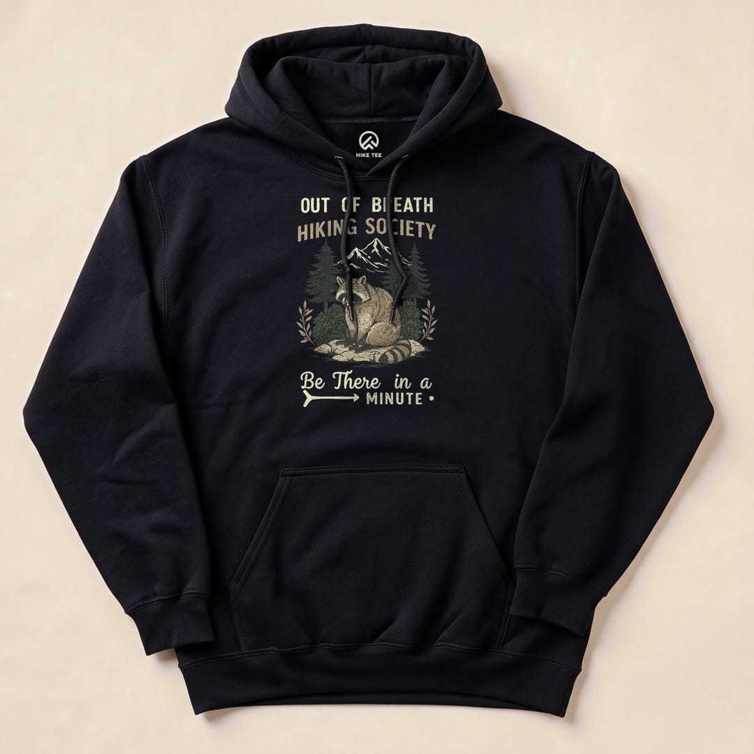 Graphic black hoodie featuring a raccoon with mountains and trees, displaying the text "Out of Breath Hiking Society" and "Be There in a Minute.