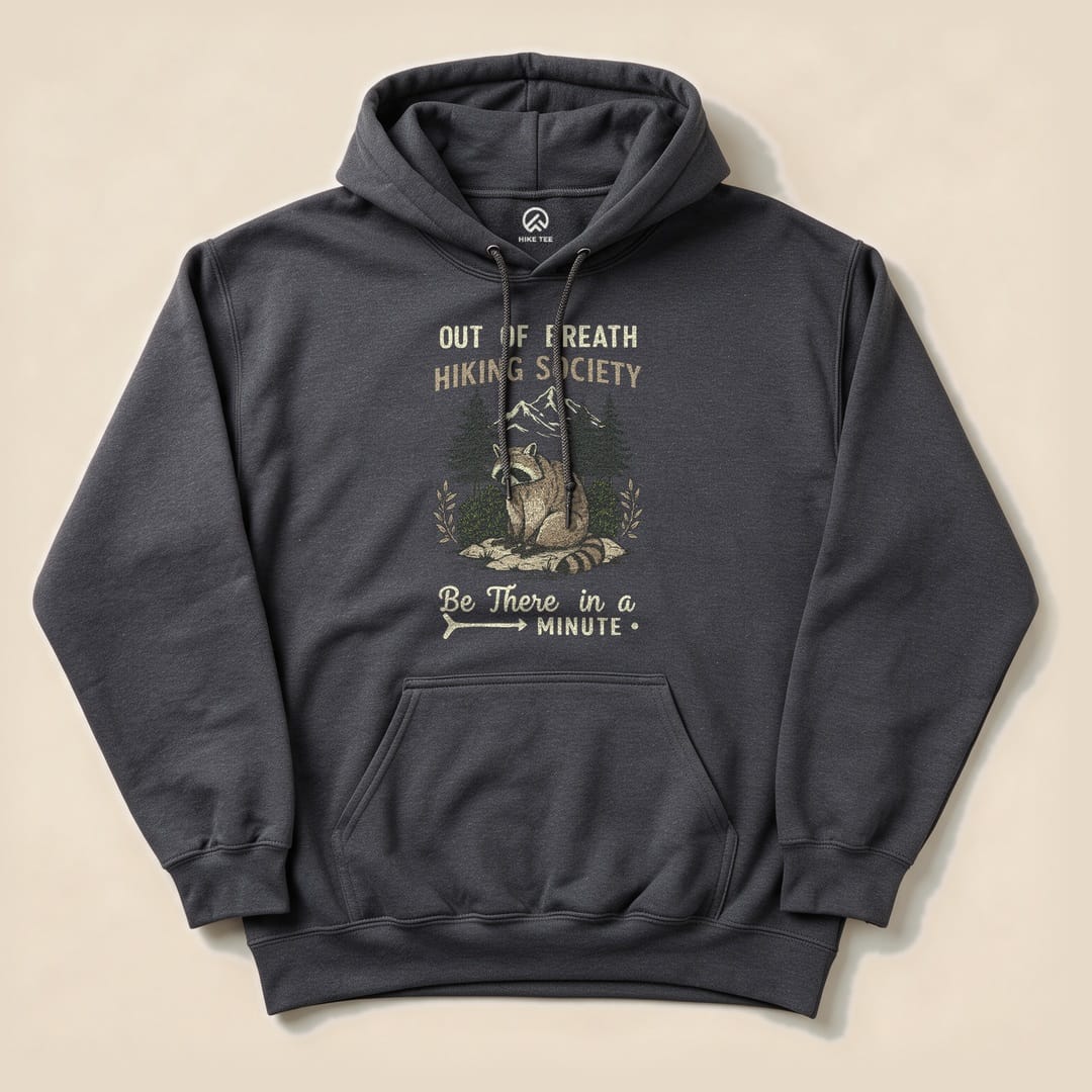 A dark hoodie featuring the "Out of Breath Hiking Society Raccoon Hoodie" graphic with a raccoon and mountains, along with the text "Be There in a Minute.