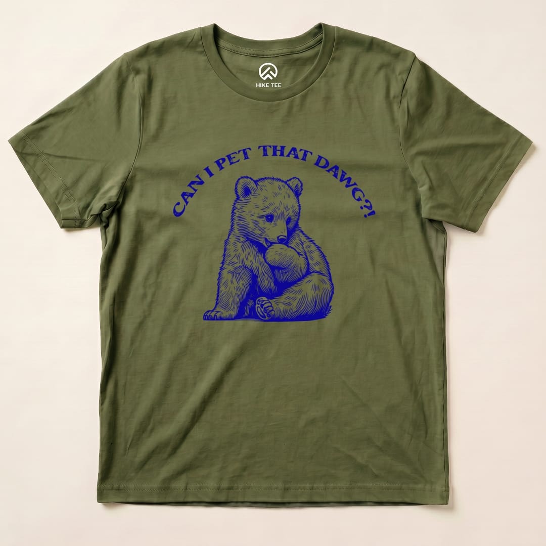 A graphic t-shirt featuring a playful bear illustration and the text "CAN I PET THAT DAWG?!" on an olive green background.
