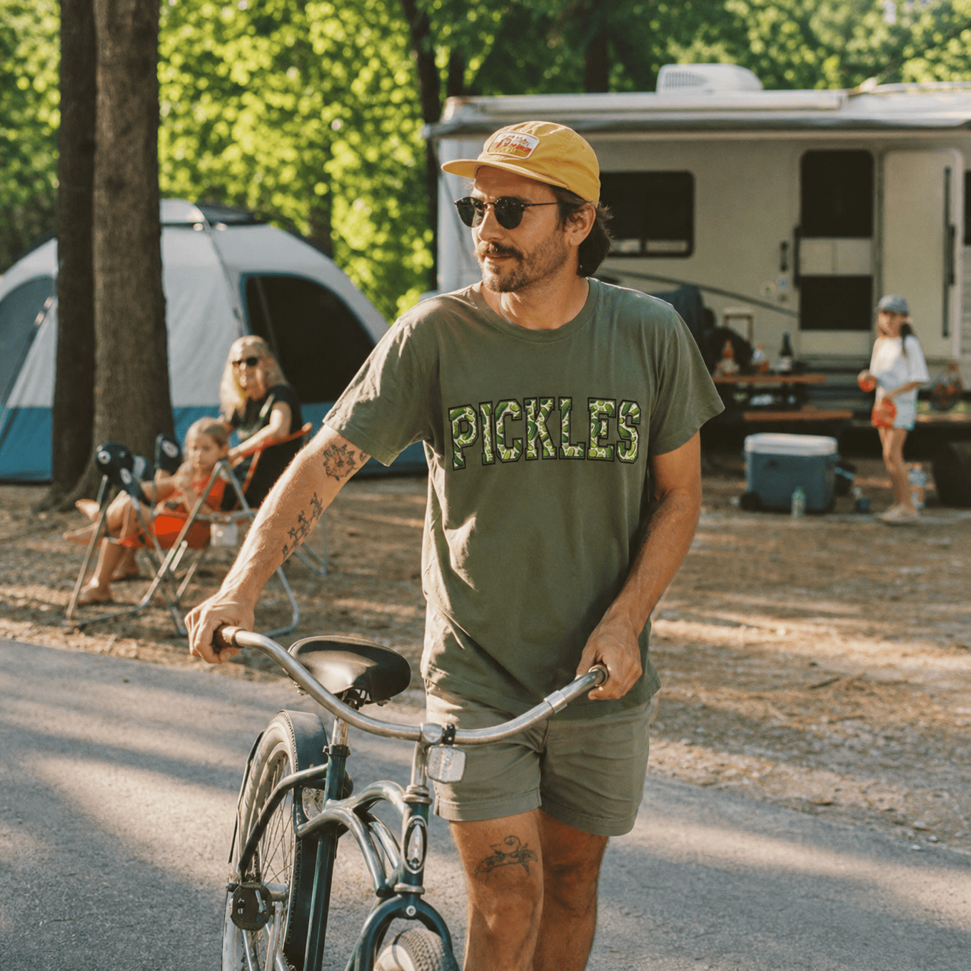 A man wears a green graphic t-shirt with the word "PICKLES" in bold letters while riding a bicycle at a campsite.
