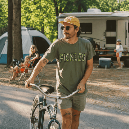 A man wears a green graphic t-shirt with the word "PICKLES" in bold letters while riding a bicycle at a campsite.