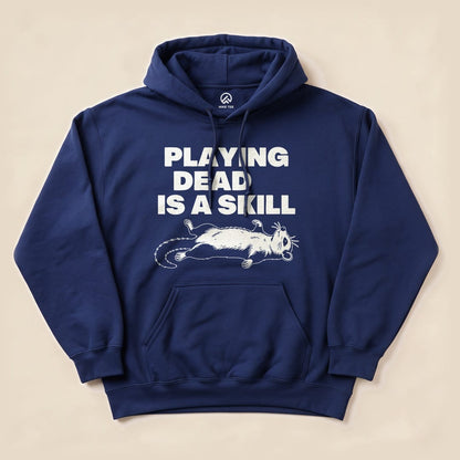 A navy hoodie features the graphic "PLAYING DEAD IS A SKILL" above an illustration of a cartoon possum lying on its back, part of the Playing Dead Possum Hoodie design.