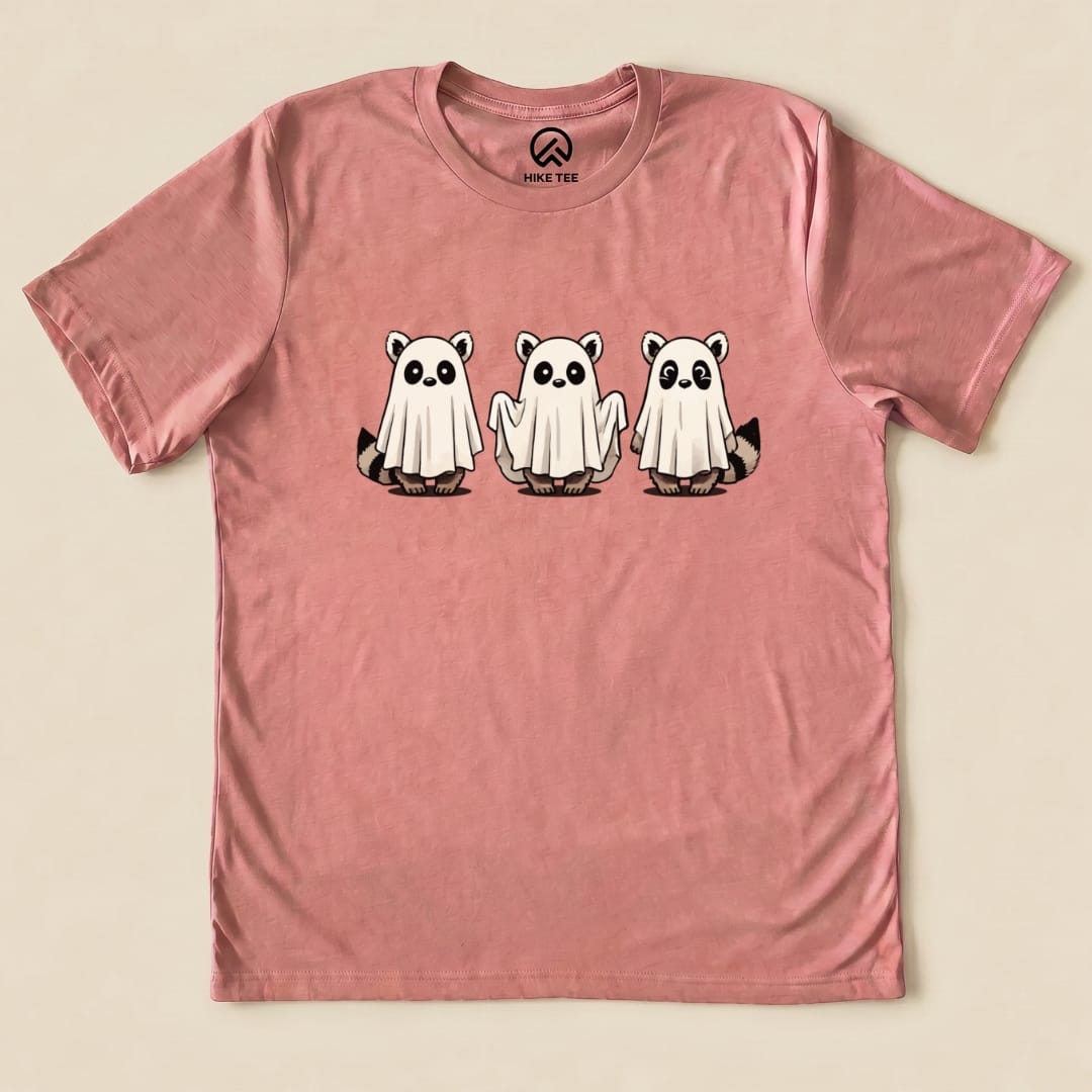 A pink graphic t-shirt featuring three raccoons wearing ghost costumes, designed as the "Raccoon Ghosts Shirt" from Hike Tee.