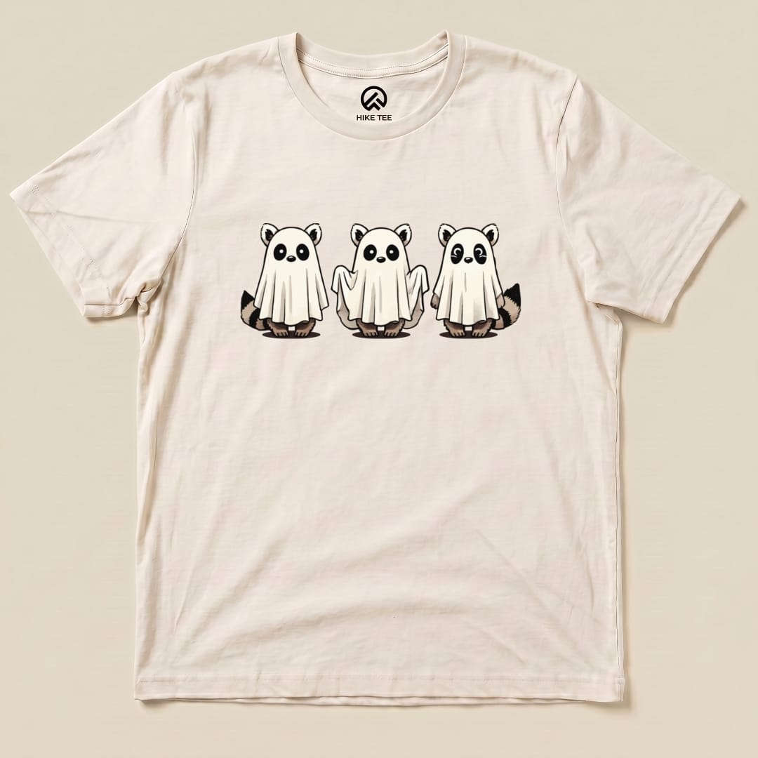 A graphic t-shirt featuring three cute raccoons dressed as ghosts, printed on a light-colored fabric, labeled as the "Raccoon Ghosts Shirt.