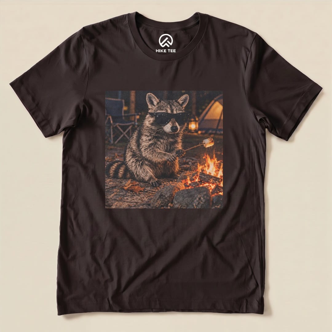 A graphic t-shirt featuring a raccoon wearing sunglasses while roasting marshmallows over a campfire, titled "Raccoon in Sunglasses Shirt.
