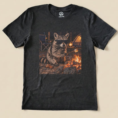 Graphic t-shirt featuring a raccoon in sunglasses, roasting a marshmallow over a campfire in a forest setting.