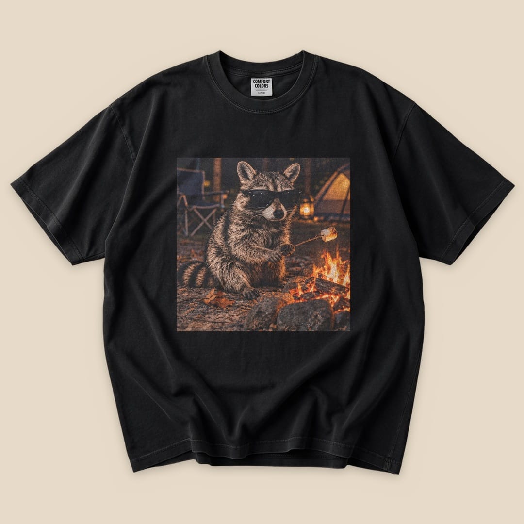 A black graphic t-shirt featuring a raccoon in sunglasses sitting by a campfire, holding a marshmallow on a stick.