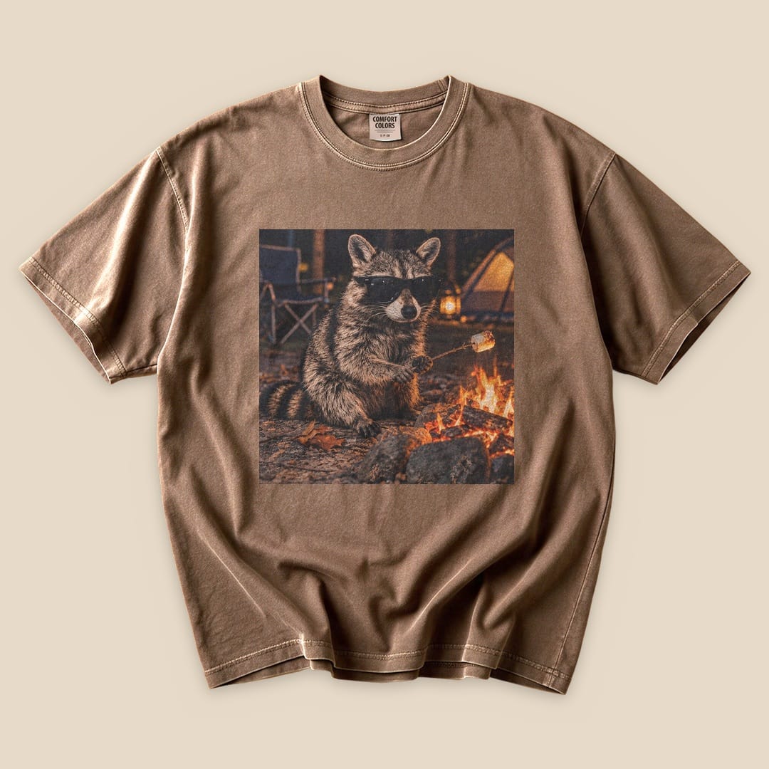 A graphic t-shirt featuring a raccoon in sunglasses sitting by a campfire, printed on a brown tee.