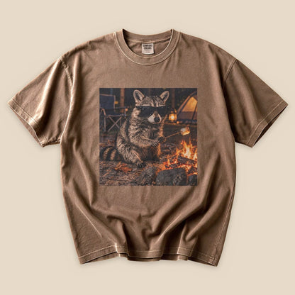 A graphic t-shirt featuring a raccoon in sunglasses sitting by a campfire, printed on a brown tee.