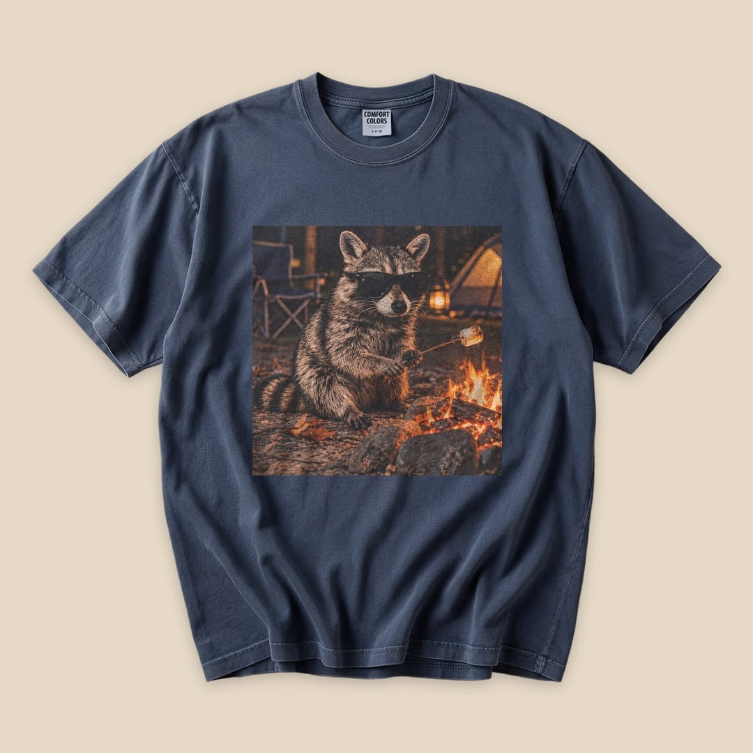A graphic t-shirt featuring a raccoon in sunglasses sitting by a campfire, capturing a humorous outdoor vibe.