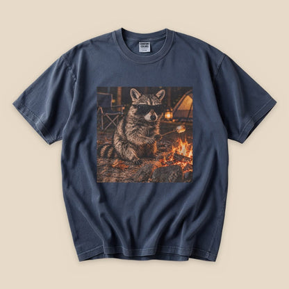 A graphic t-shirt featuring a raccoon in sunglasses sitting by a campfire, capturing a humorous outdoor vibe.
