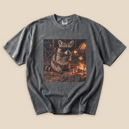 A graphic t-shirt featuring a raccoon in sunglasses sitting by a campfire, showcasing a playful outdoor vibe.