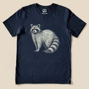 A navy graphic t-shirt features an illustration of a raccoon with a humorous expression, labeled as the "Raccoon Is Watching You Shirt.