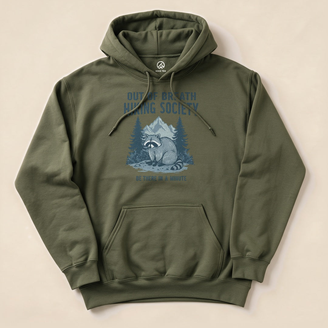 Graphic t-shirt featuring a raccoon sitting in front of trees and mountains with the phrase "Out of Breath Hiking Society" and "Be There In A Minute.