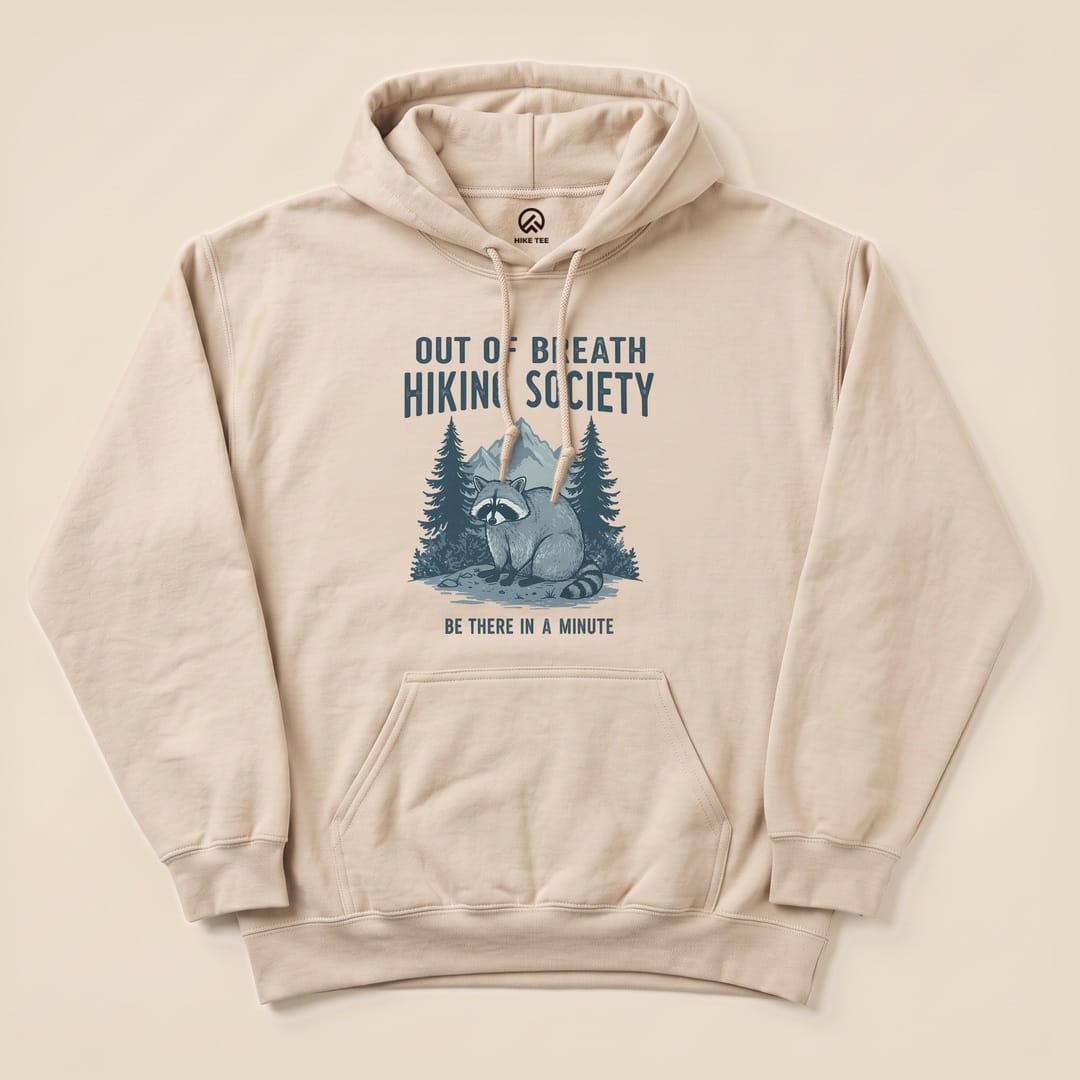 Beige "Raccoon Out Of Breath Hoodie" featuring a raccoon illustration amidst trees, with the text "Out of Breath Hiking Society" and "Be There in a Minute".