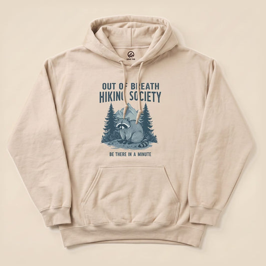 Beige "Raccoon Out Of Breath Hoodie" featuring a raccoon illustration amidst trees, with the text "Out of Breath Hiking Society" and "Be There in a Minute".