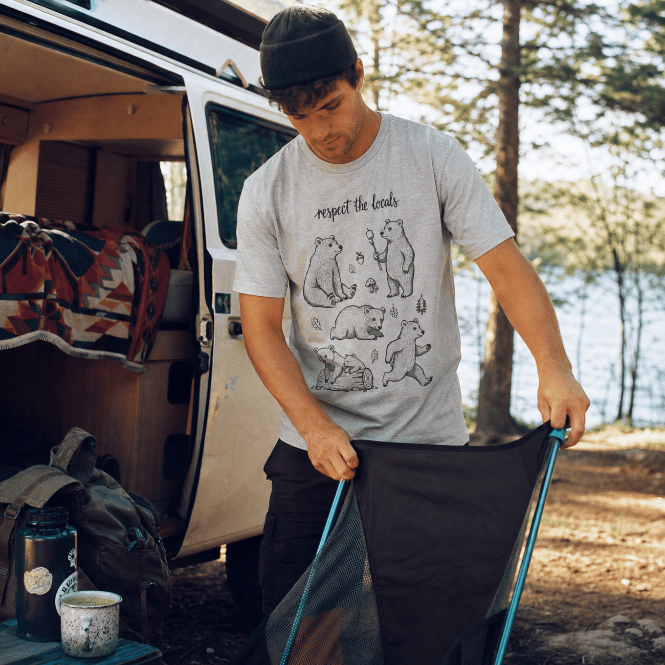 A man outdoors near a camper van is wearing a graphic t-shirt featuring playful bear illustrations and the phrase "respect the locals.