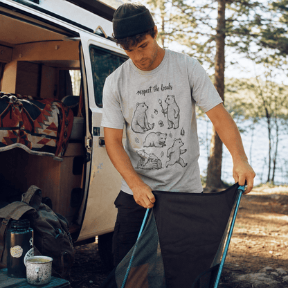 A man outdoors near a camper van is wearing a graphic t-shirt featuring playful bear illustrations and the phrase "respect the locals.