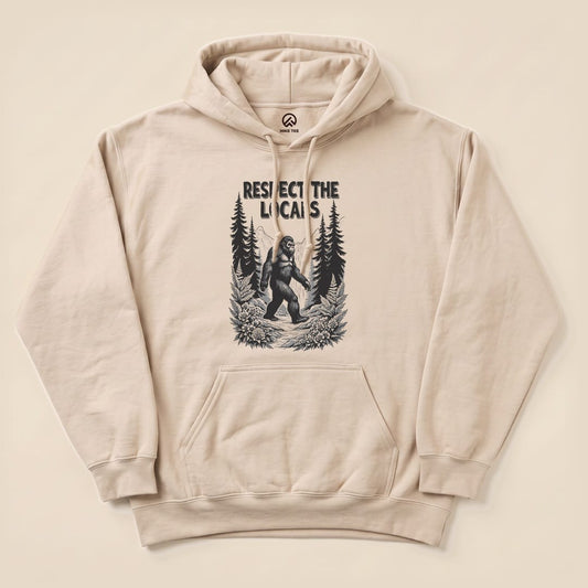 A beige hoodie features a graphic of Bigfoot walking through a forest with the text "Respect The Locals Bigfoot Hoodie".