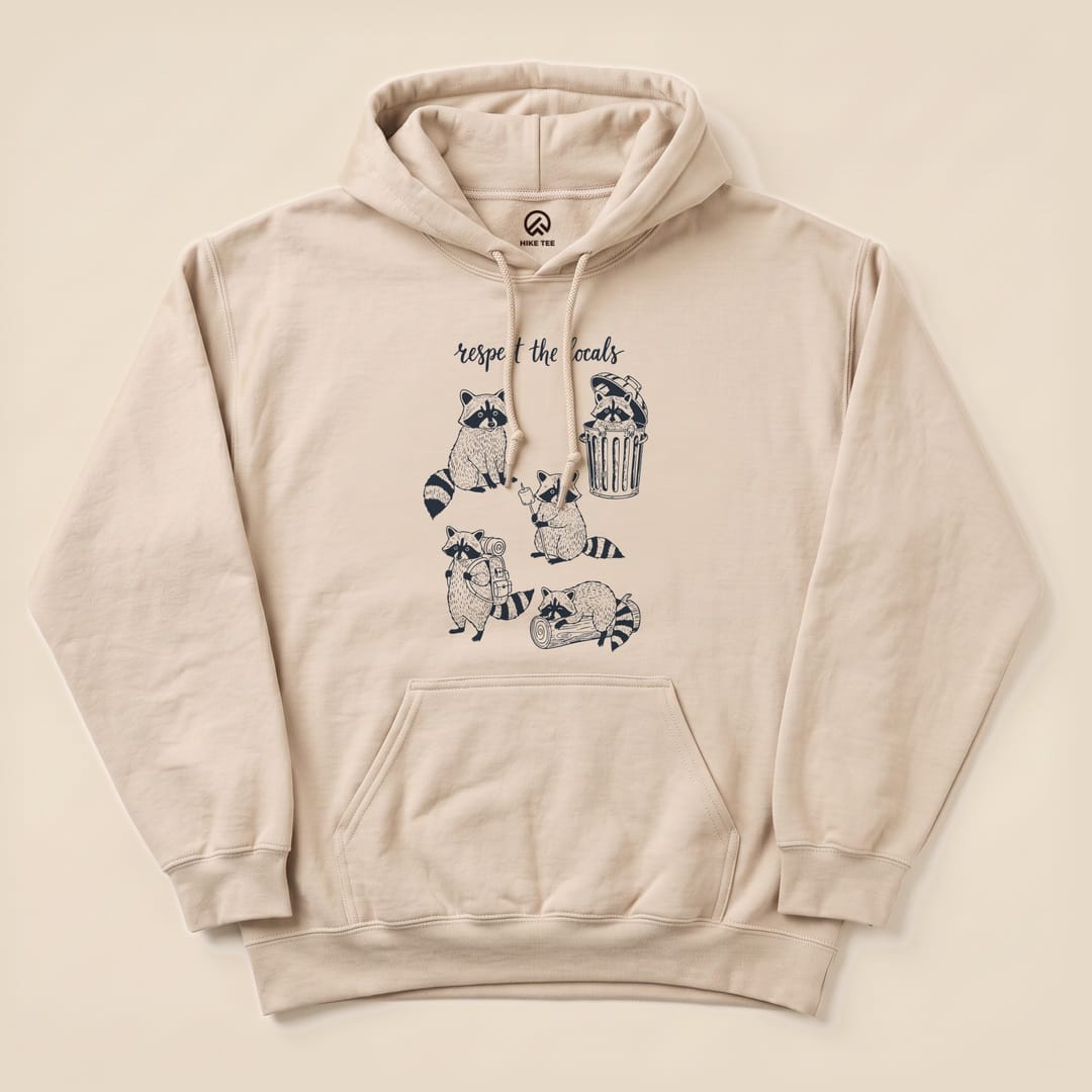 The “Respect the Raccoons Hoodie” features a playful graphic of raccoons engaging in quirky activities, printed on a beige hoodie.