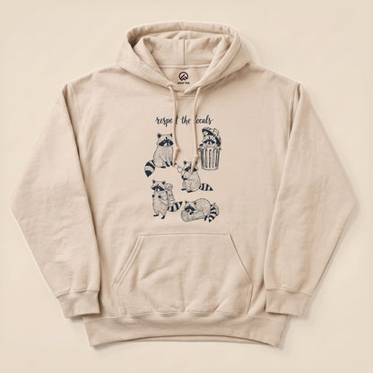 The “Respect the Raccoons Hoodie” features a playful graphic of raccoons engaging in quirky activities, printed on a beige hoodie.